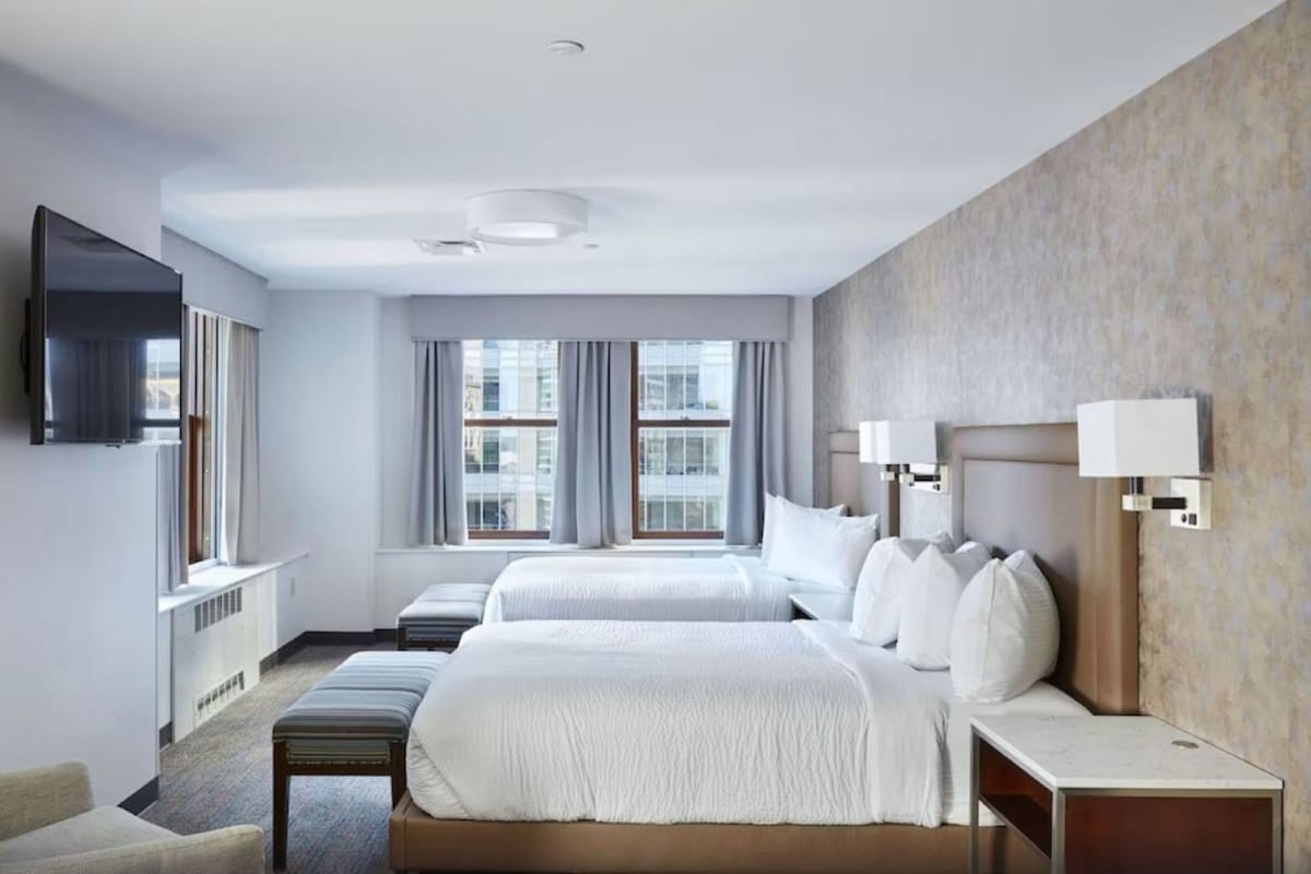 Habitación de hotel con dos camas, un televisor de pantalla plana, un escritorio con lámpara y vistas a la ciudad a través de amplias ventanas.
