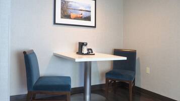 Una habitación pequeña con una mesa blanca y dos sillas azules, un cuadro en la pared y una cafetera sobre la mesa.