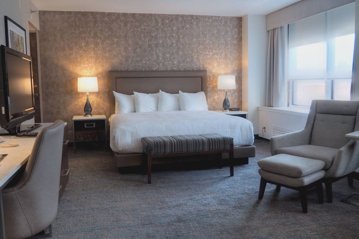 A hotel room with a large bed, two bedside lamps, a desk with a computer, a chair, and a TV.