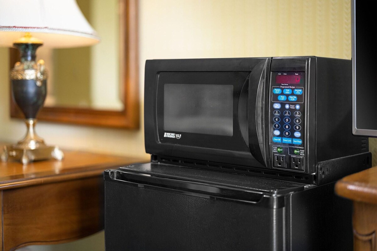 A microwave oven with a digital interface, placed on a black appliance, likely a refrigerator, in a hotel room.