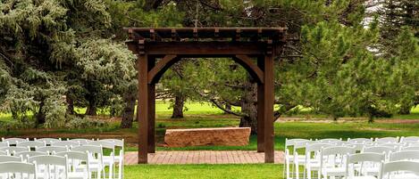 A gazebo with a brick base and wooden beams, surrounded by white chairs arranged in rows on a grassy lawn, with trees in the background.
