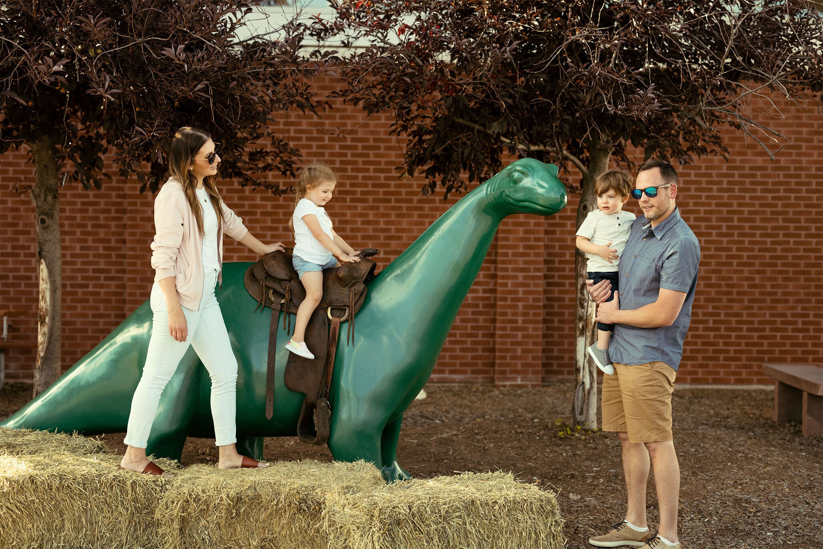 A family interacting with a green dinosaur sculpture in an outdoor setting with a brick wall and trees in the background.