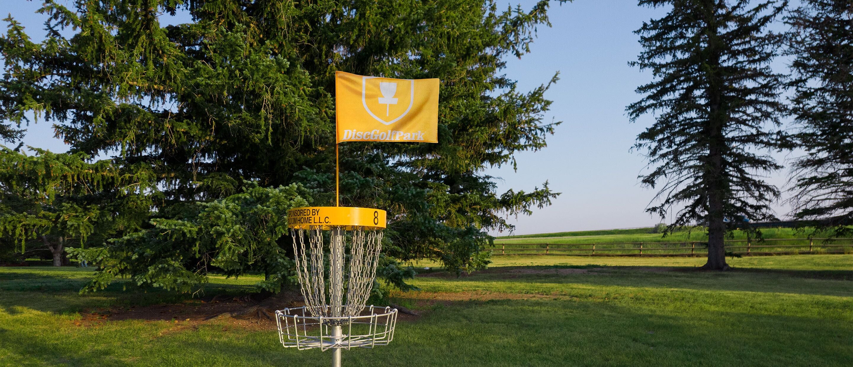 A disc golf basket with a yellow flag and a logo, surrounded by trees and a grassy field.