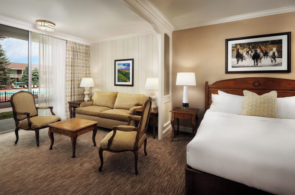 A hotel room with a bed, a sofa, two armchairs, a small table, and a nightstand with lamps. There is a framed picture on the wall and a window with curtains.