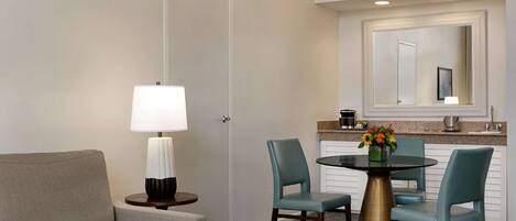 A hotel room with a sofa, a round table with chairs, a lamp, a mirror, and a kitchen area with a countertop and sink.