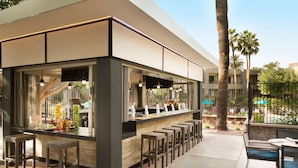 An outdoor bar area with wicker stools, a counter with a glass display, and a view of palm trees and a pool.