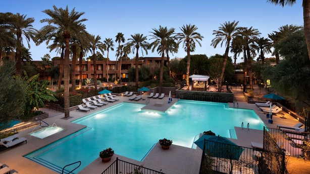A swimming pool with sun loungers and palm trees, surrounded by a building with multiple floors and balconies.
