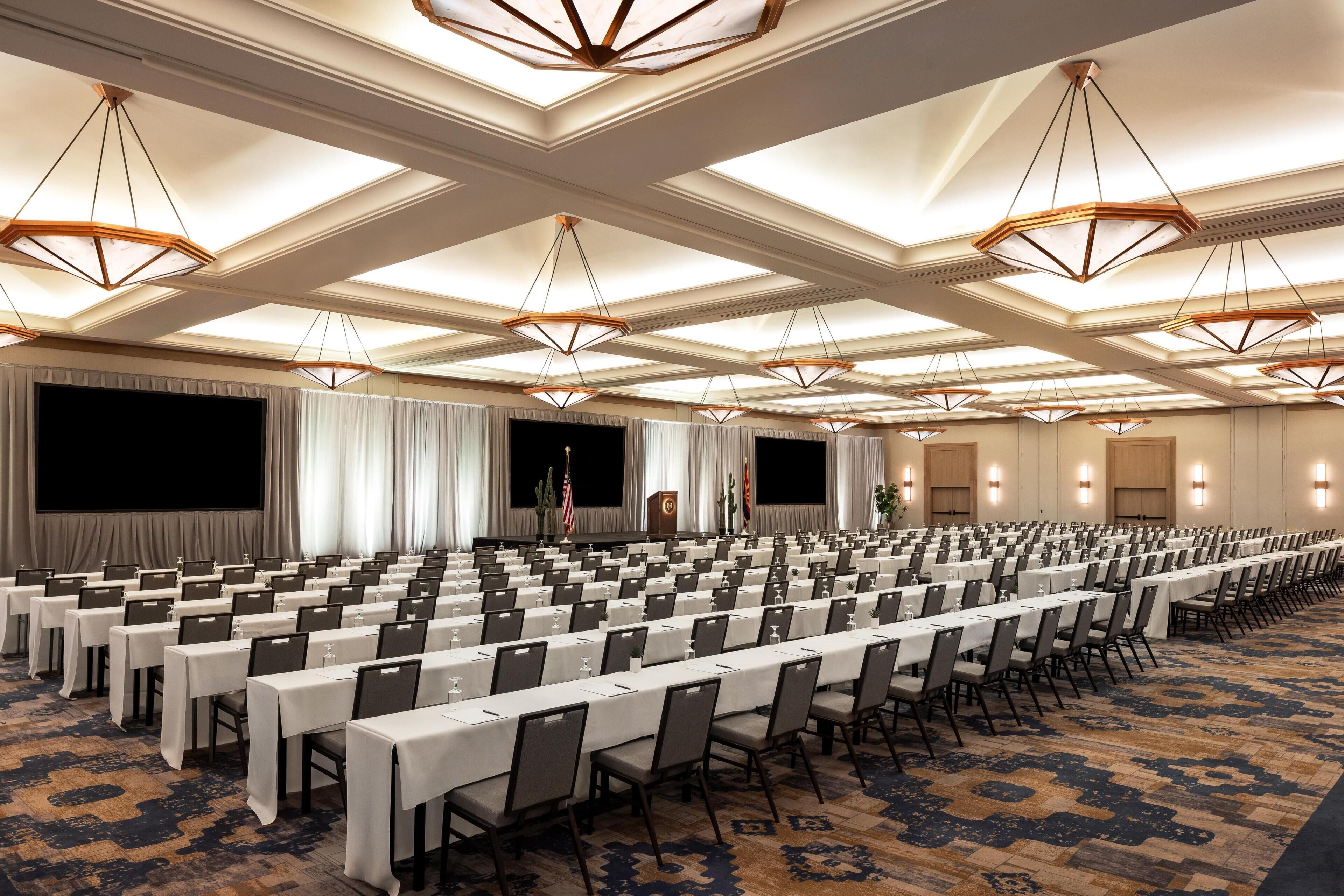 A conference room with rows of tables and chairs, a stage with curtains, and a ceiling with geometric light fixtures.