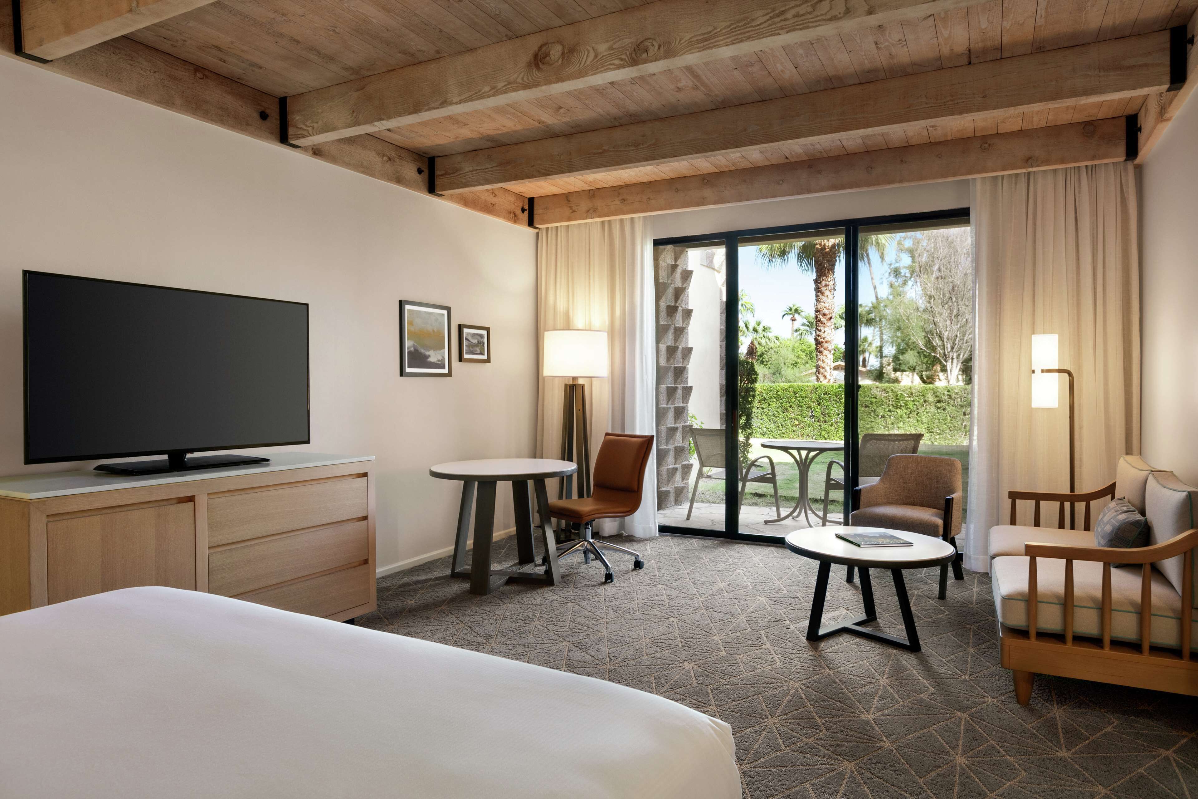 A hotel room with a bed, a TV, a desk with a chair, a small table, and a sofa. There is a large window with a view of greenery and a palm tree.
