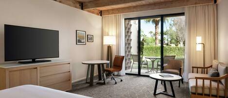 A hotel room with a bed, a TV, a desk with a chair, a small table, and a sofa. There is a large window with a view of greenery and a palm tree.