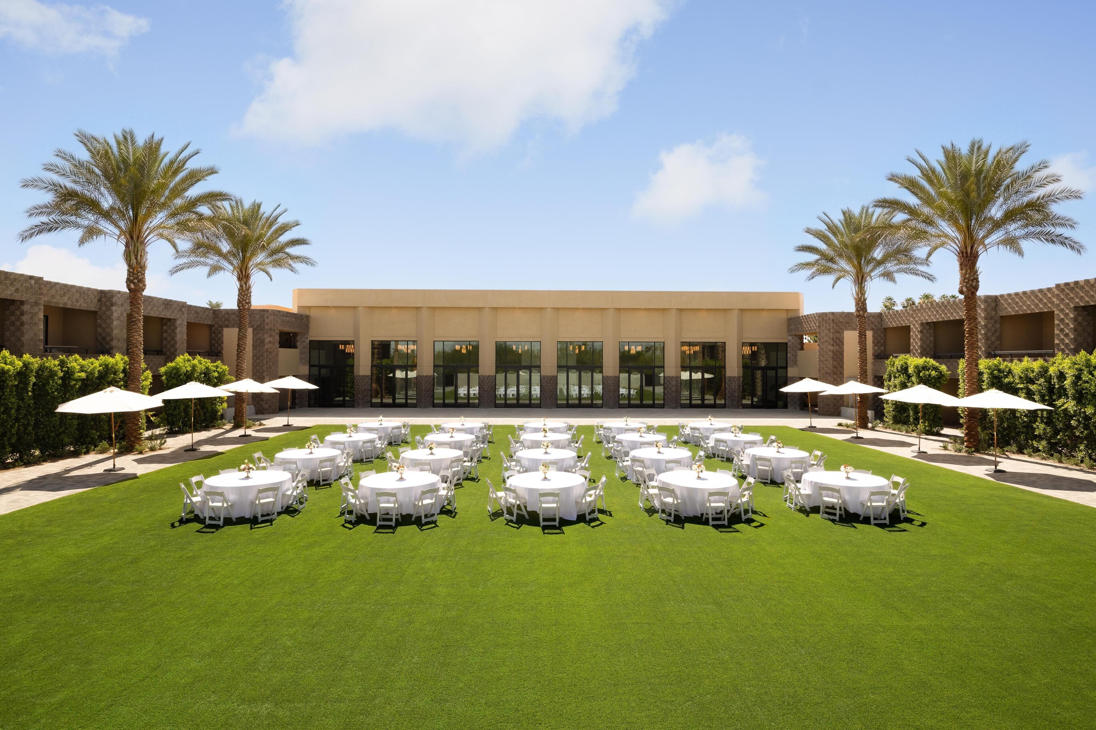 A hotel courtyard with multiple round tables set up for dining, surrounded by palm trees and a building with large windows.
