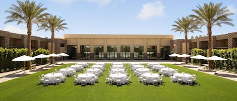 A hotel courtyard with multiple round tables set up for dining, surrounded by palm trees and a building with large windows.