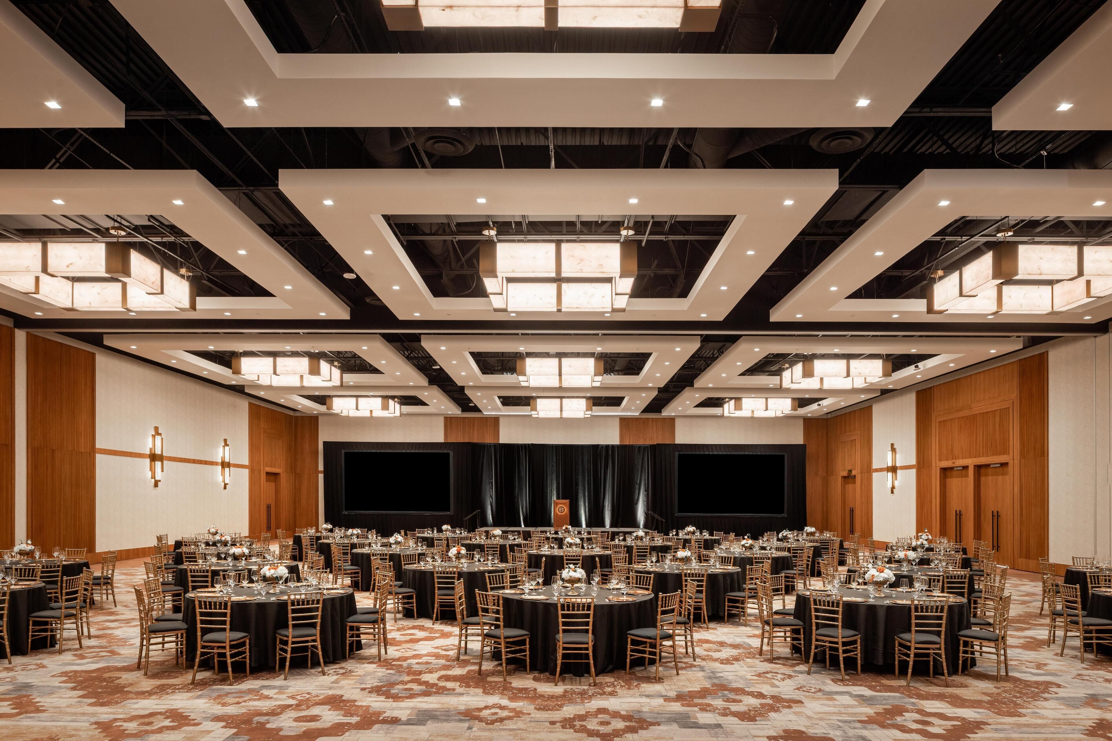 A large banquet hall with multiple round tables, black tablecloths, and gold chairs.