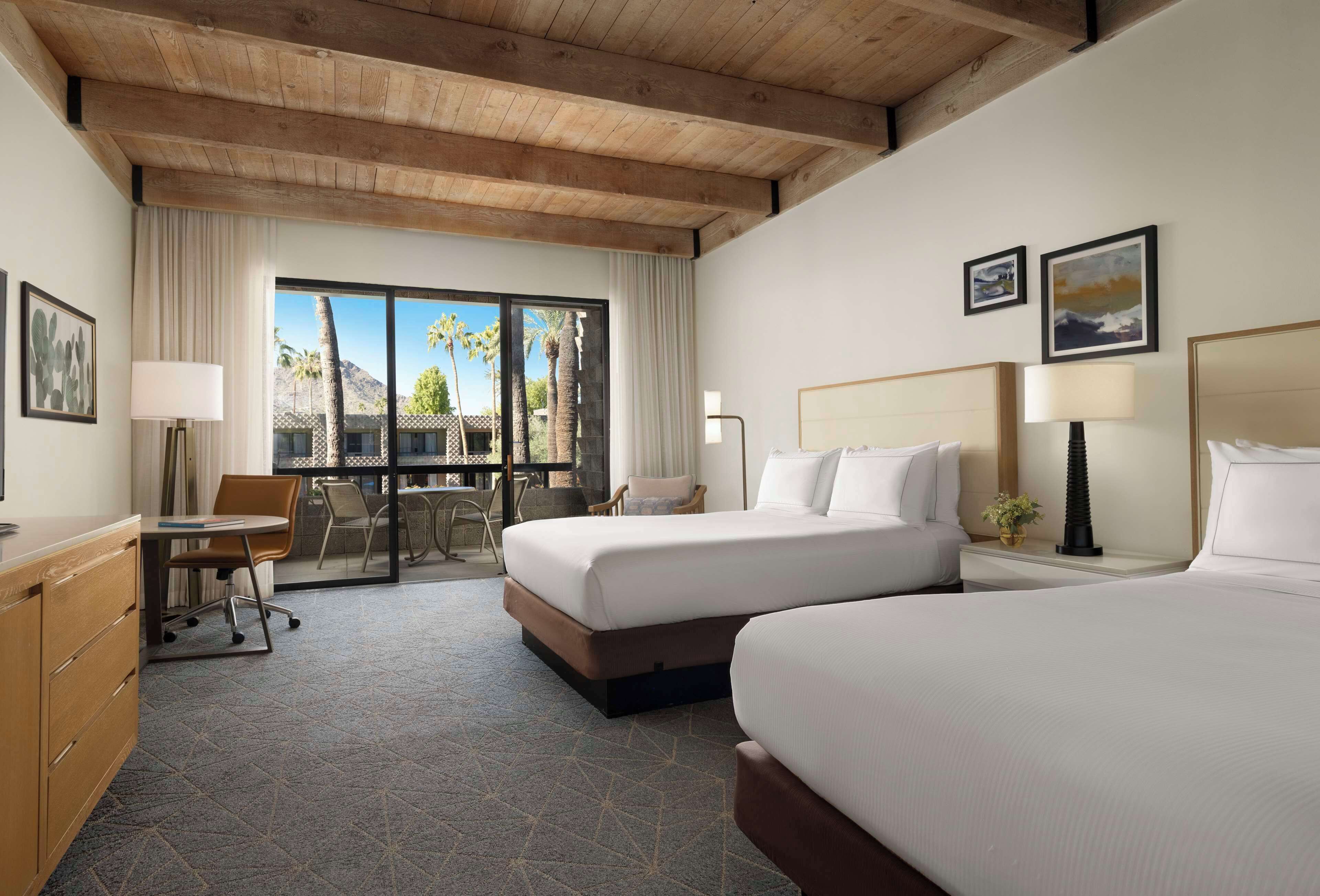 A hotel room with a large bed, a desk, a chair, and a view of palm trees through the window.