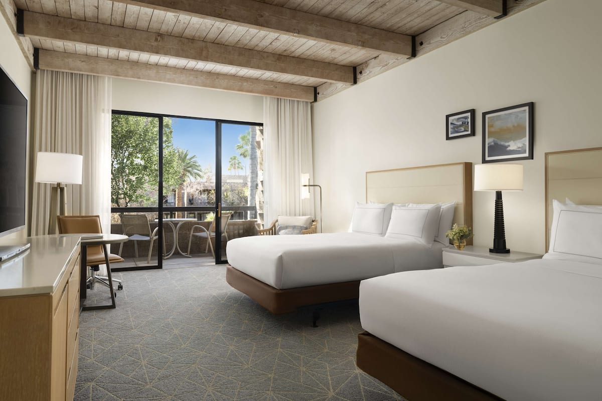 Une chambre d’hôtel avec deux lits, un bureau, une chaise, un balcon donnant sur des palmiers et une grande fenêtre.