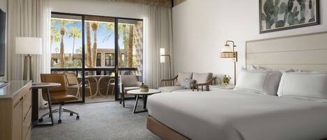 A hotel room with a large bed, a desk, a chair, a small table, and a balcony with a view of palm trees and buildings.