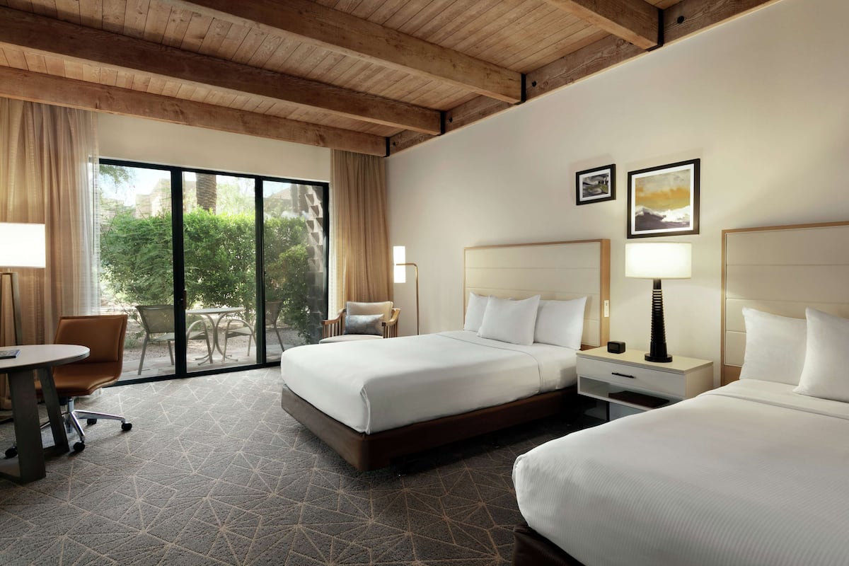 Une chambre d’hôtel avec deux lits, un bureau et une chaise. Une grande fenêtre offre une vue sur la verdure environnante.