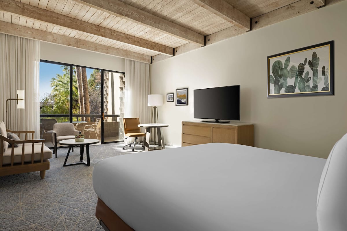Une chambre d’hôtel avec un grand lit, un téléviseur à écran plat, un bureau et une chaise. Il y a une fenêtre avec des rideaux et une vue sur des palmiers.