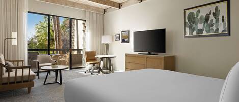Quarto de hotel com uma cama grande, televisão de tela plana, escrivaninha e uma cadeira. Há uma janela com cortinas e vista para palmeiras.