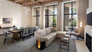 A modern hotel room with a wooden ceiling, a large sectional sofa, a dining table with chairs, a small coffee table, and large windows with curtains.