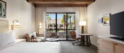 Habitación de hotel con una cama grande, un escritorio, dos sillas, un televisor y un balcón con vistas a palmeras y edificios.