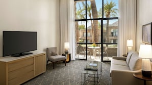 A hotel room with a large window, a flat-screen TV, a sofa, a chair, a coffee table, and a lamp.