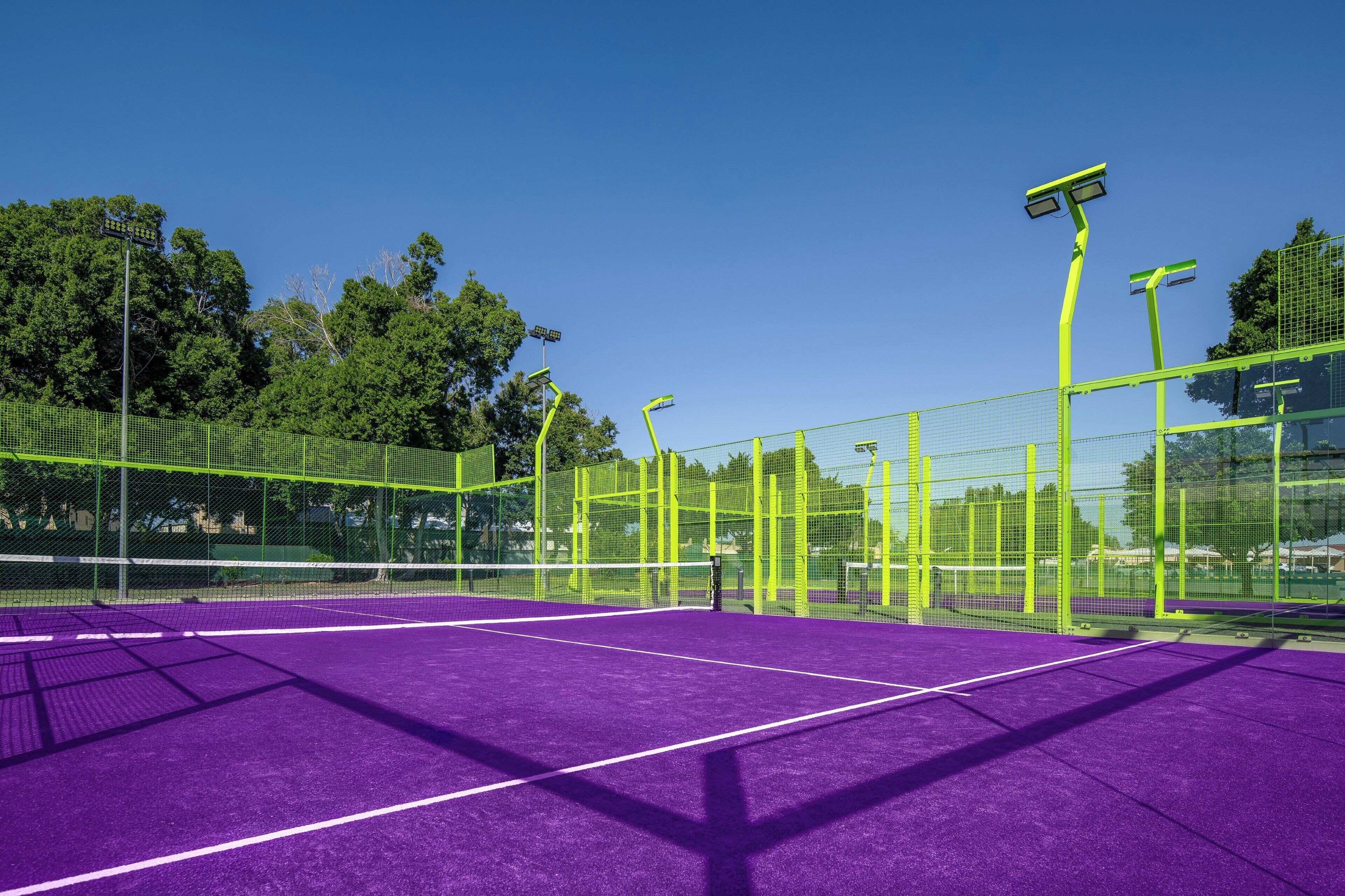 Ein Außen-Tennisplatz mit violetter Belag, umgeben von einem grünen Zaun und Netz, ausgestattet mit Flutlicht.