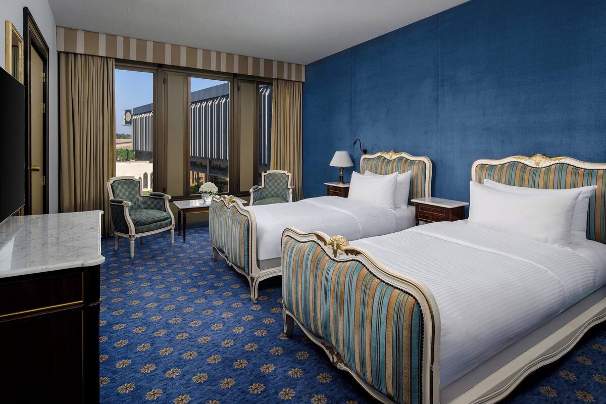 A hotel room with two beds, a desk, a chair, and a large window with curtains.