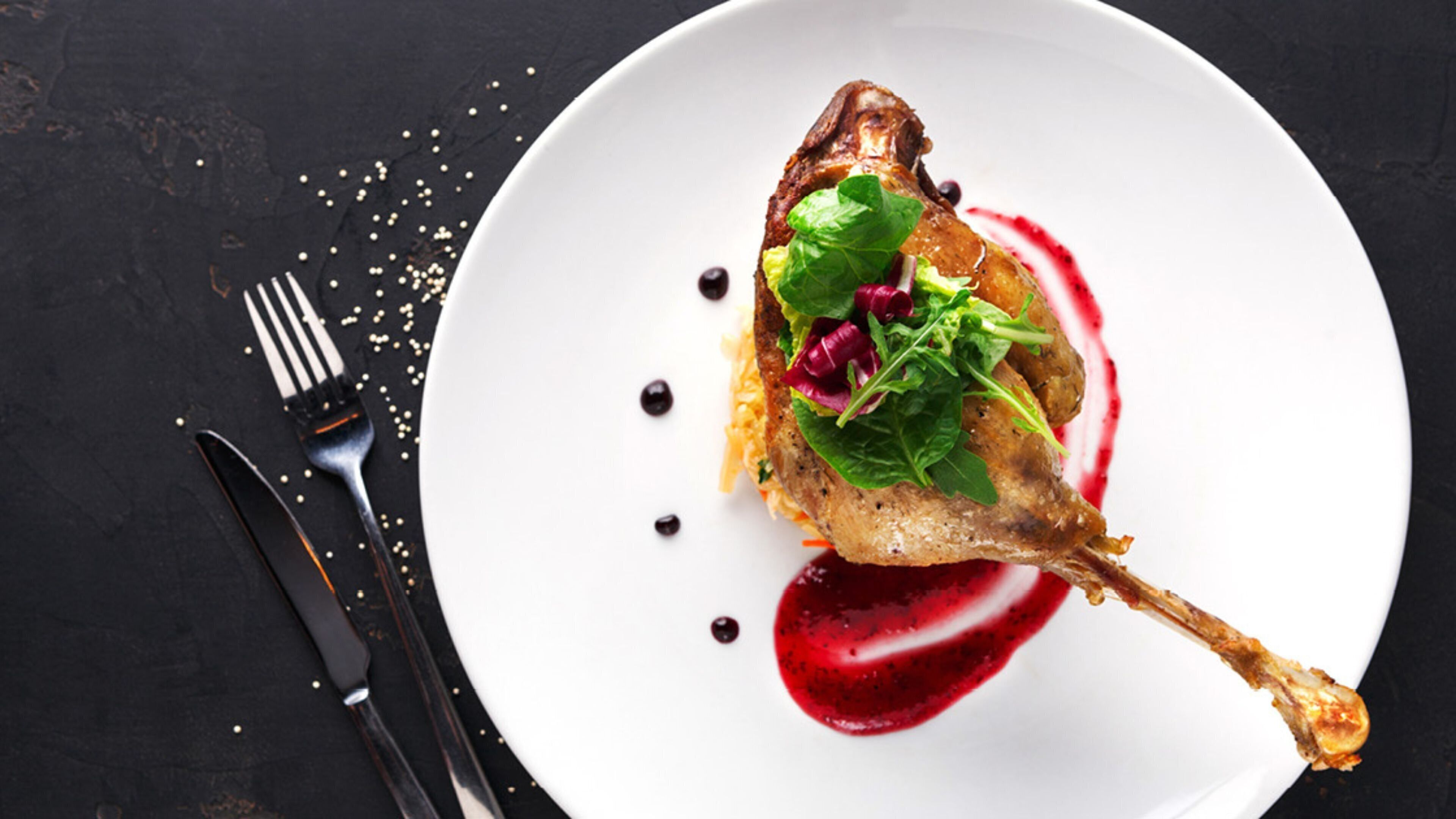 A white plate with a roasted duck leg, garnished with greens and a red sauce, on a dark surface.