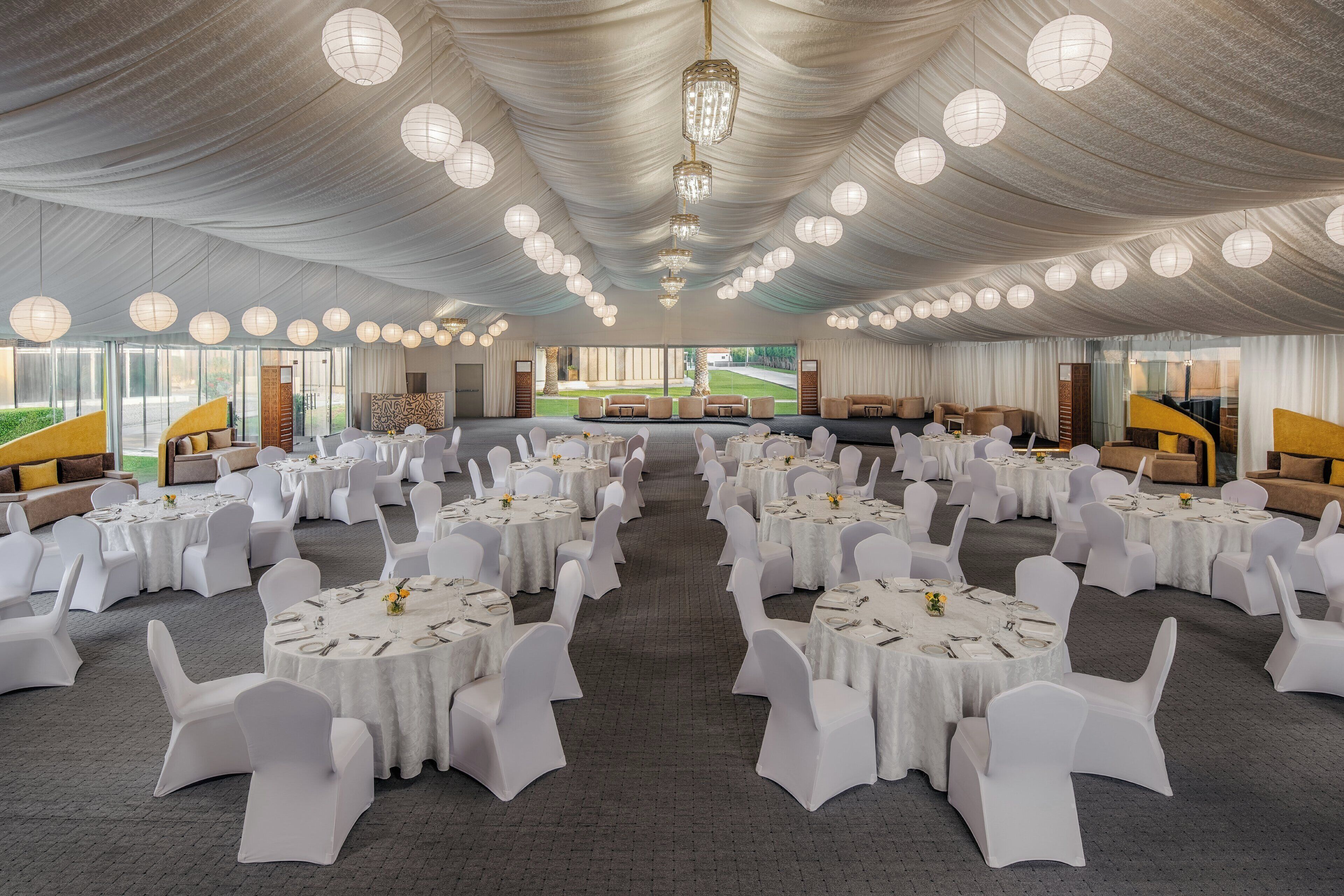 A banquet hall with round tables and white chairs, large windows, and a chandelier.