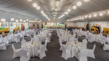 A banquet hall with round tables and white chairs, large windows, and a chandelier.