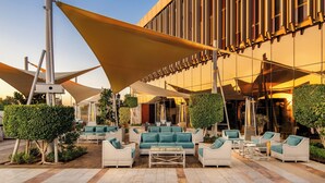 An outdoor seating area with white sofas and tables, shaded by large umbrellas, in front of a modern building with large windows.
