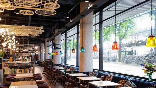 Interior de restaurante con techos altos, amplias ventanas y diversas opciones de asientos, como mesas y cabinas.