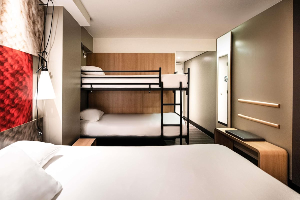A hotel room with bunk beds, a bedside table, a lamp, a mirror, and a wardrobe.