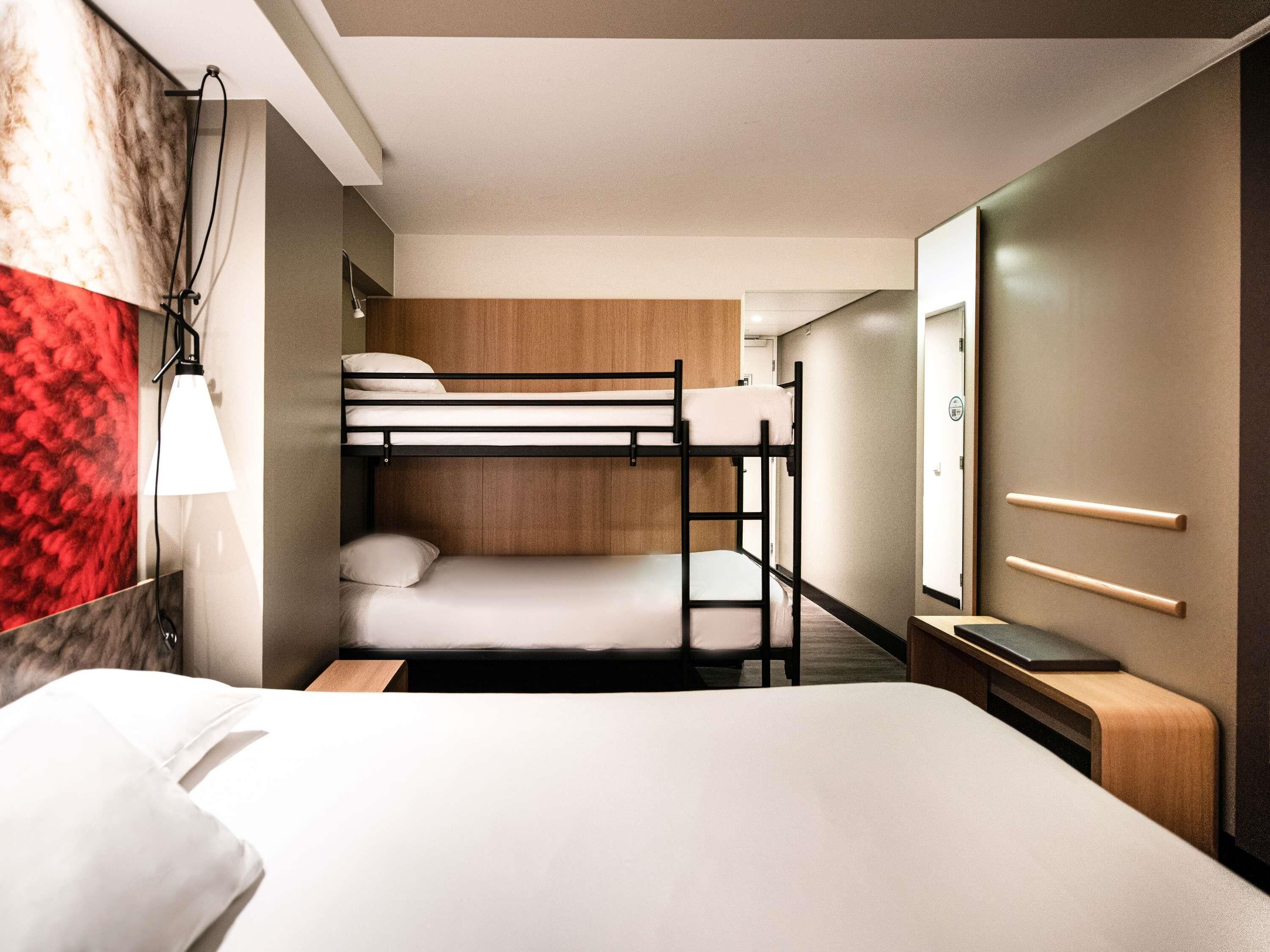 A hotel room with bunk beds, a bedside table, a lamp, a mirror, and a wardrobe.
