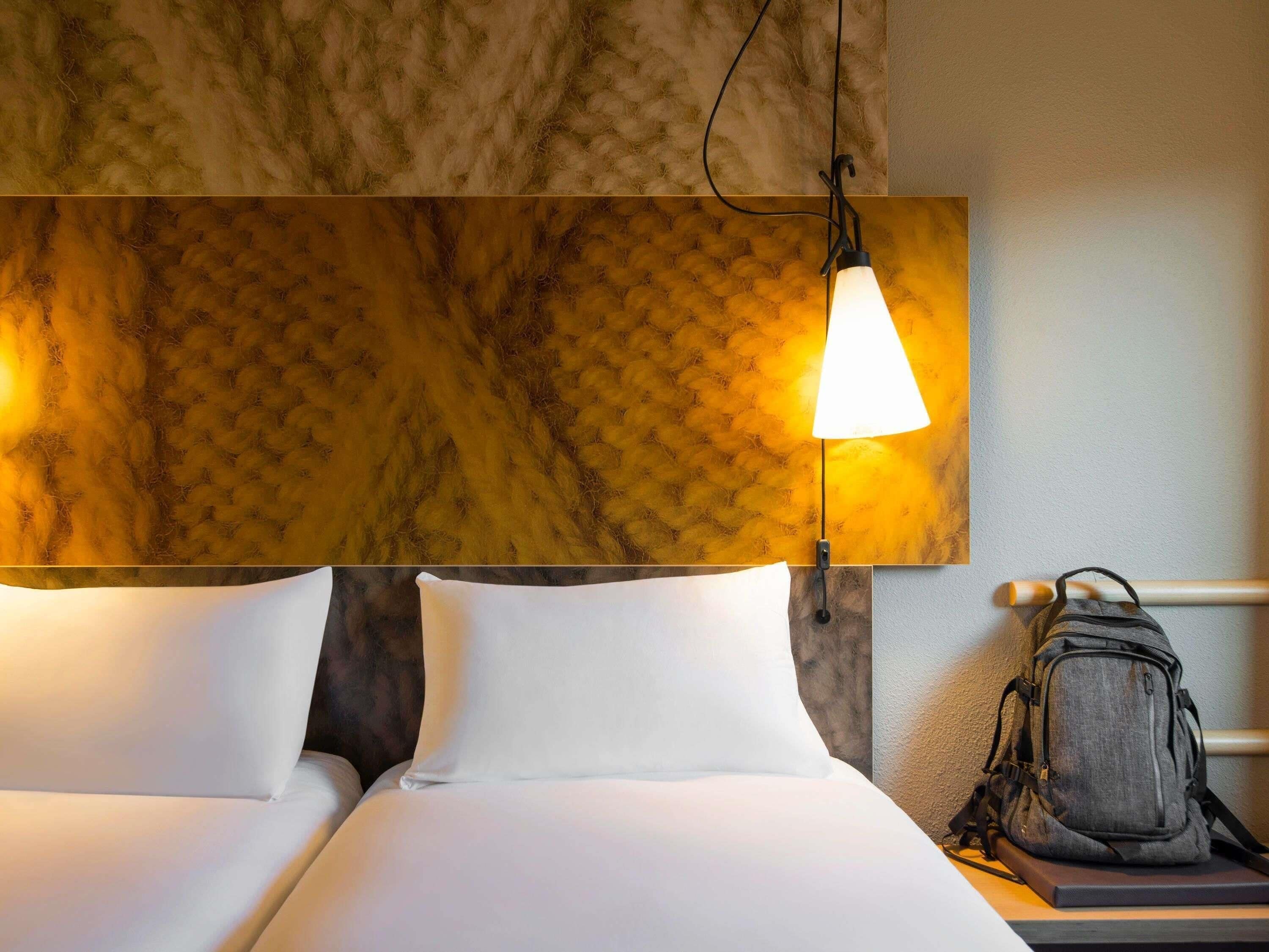 A hotel room with a bed, pillows, a lamp, a wall with a textured pattern, and a backpack on a nightstand.