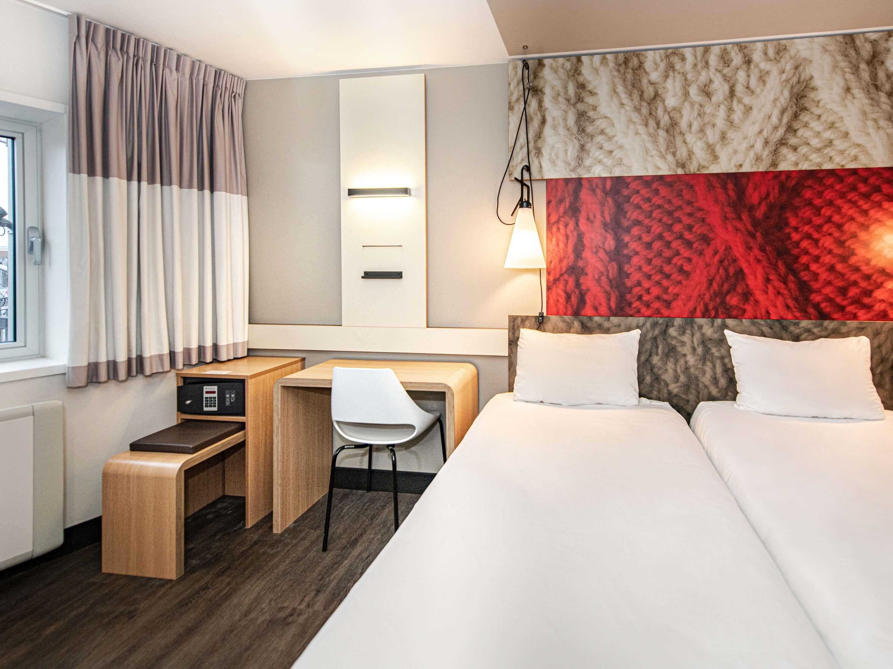 Una habitación de hotel con cama, escritorio, silla y un ventanal con cortinas.