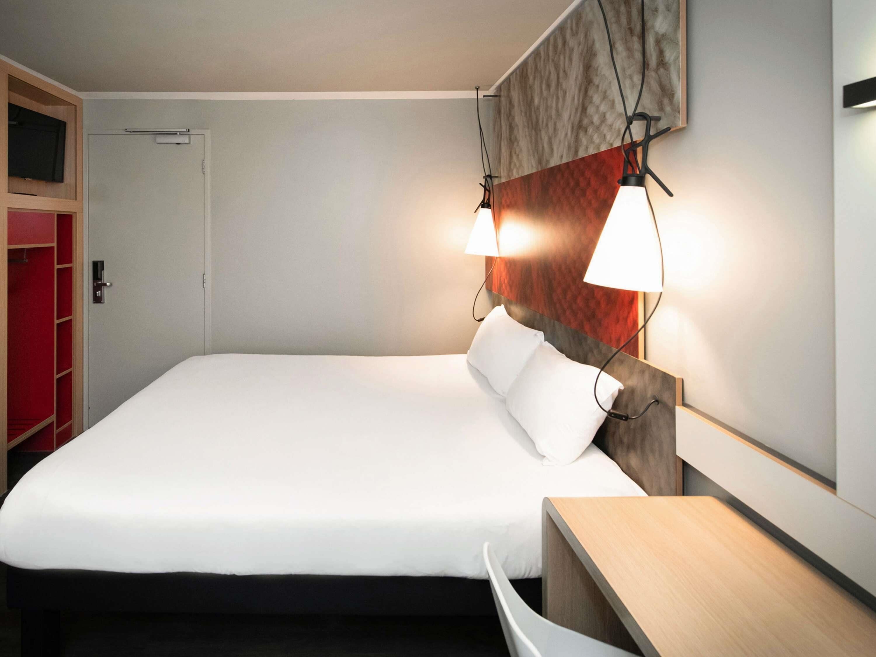 A neatly made bed with white linens, a wooden bedside table, and a wall-mounted lamp.