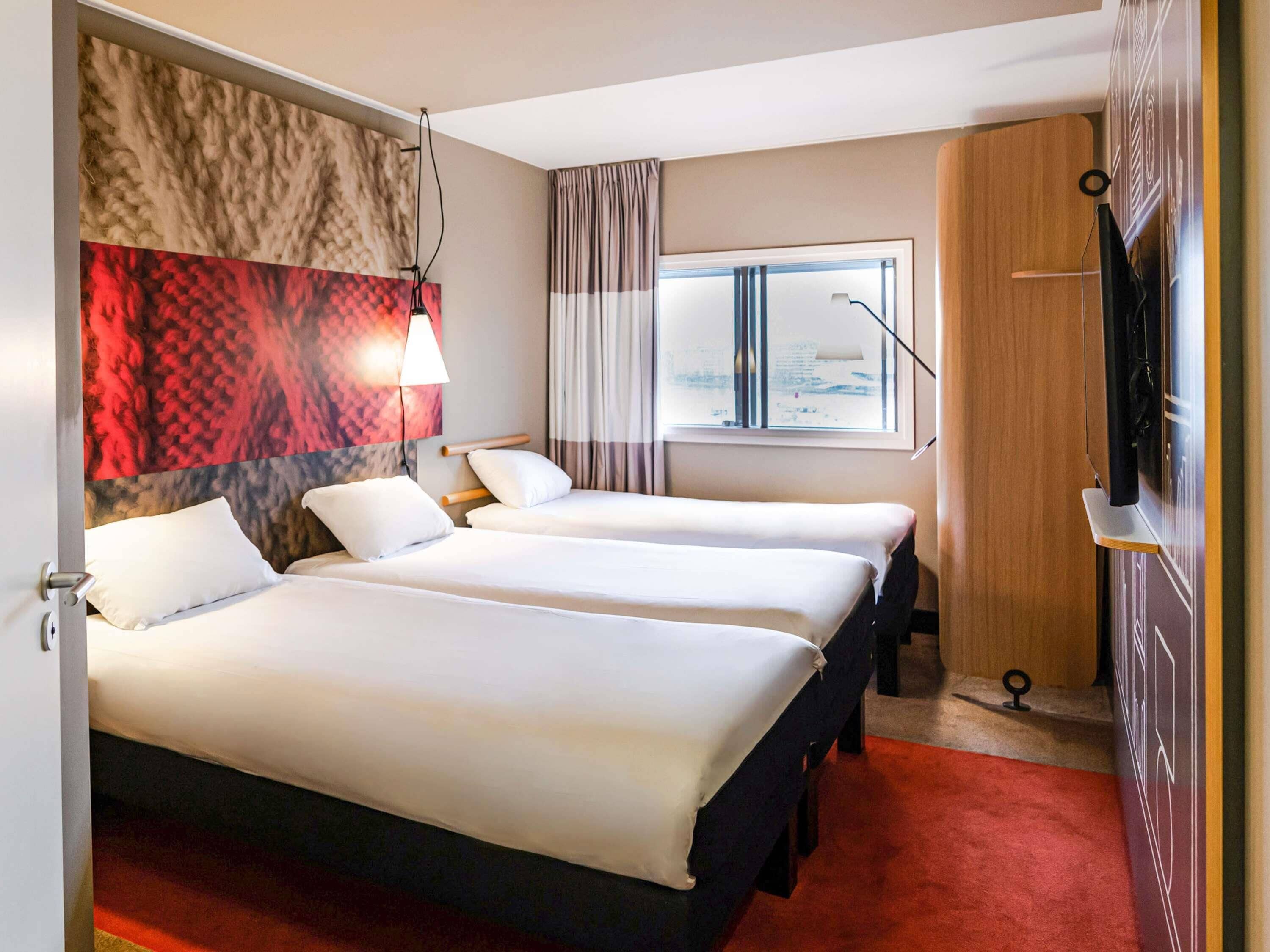 A hotel room with two beds, a large window, a TV, and a red carpet.