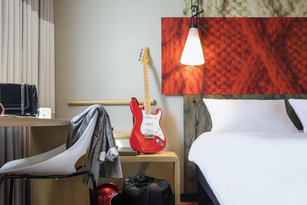 Une chambre d’hôtel avec un lit, un bureau sur lequel se trouve une guitare, une chaise, une valise et un casque rouge.