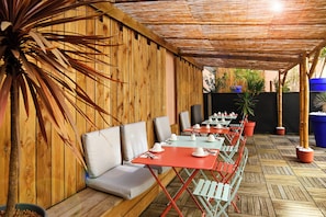 An outdoor seating area with wooden walls, cushioned benches, and tables set for dining.