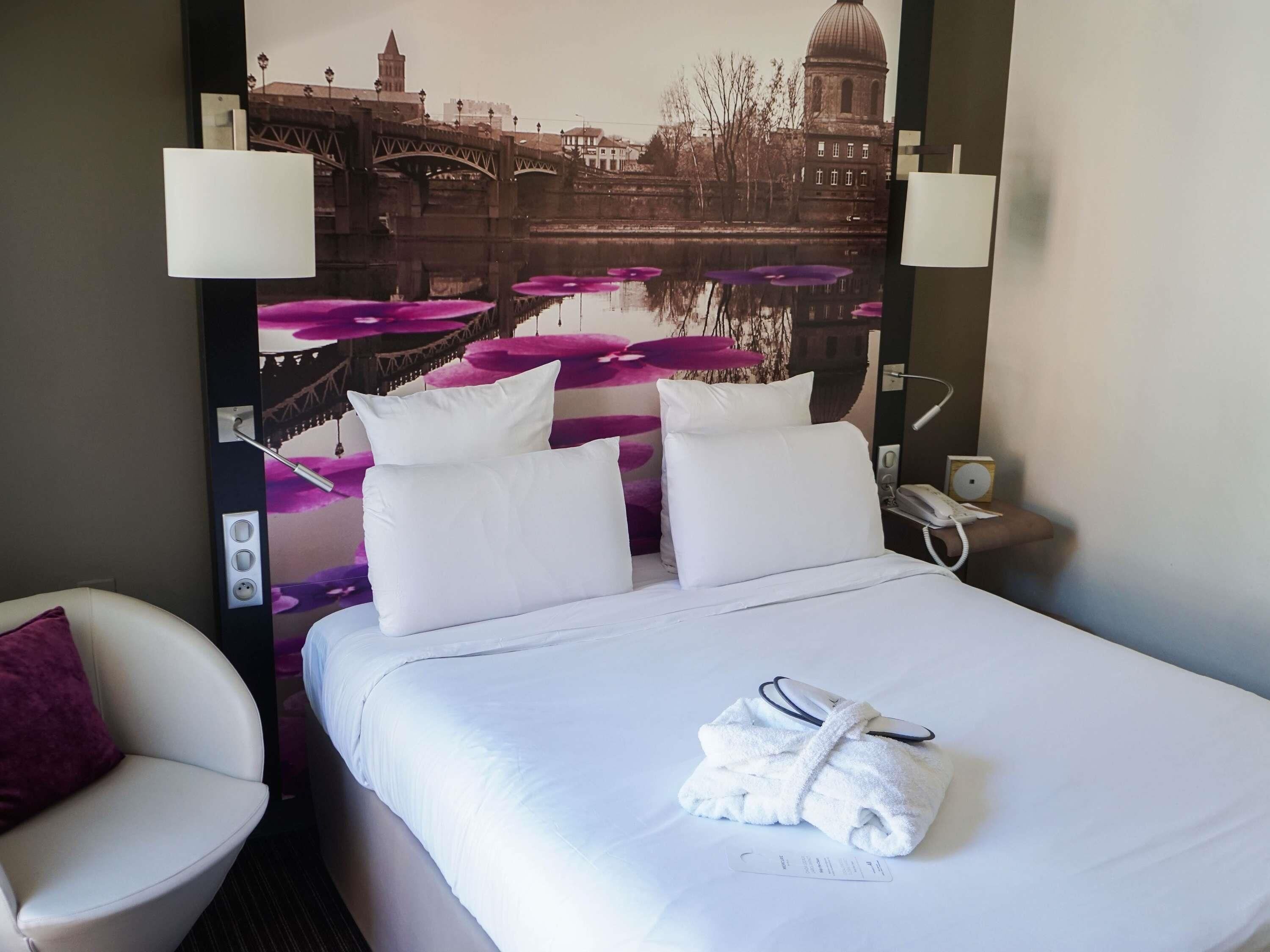 Habitación de hotel con cama, dos almohadas blancas, bata blanca, mesita de noche con teléfono y lámpara de pared.