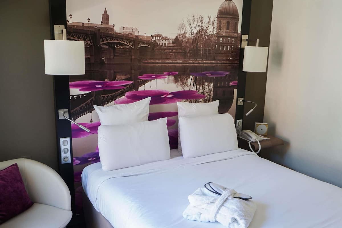 Habitación de hotel con cama, dos almohadas blancas, bata blanca, mesita de noche con teléfono y lámpara de pared.
