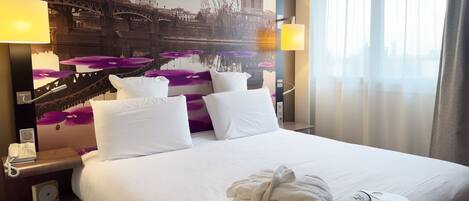 A hotel room with a large bed, white bedding, a robe, and a remote control on the bed. There is a nightstand with a phone and a lamp. A large wall mural depicts a bridge and a building.