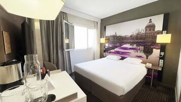 A hotel room with a bed, a desk with a lamp, a coffee maker, and a window with curtains.