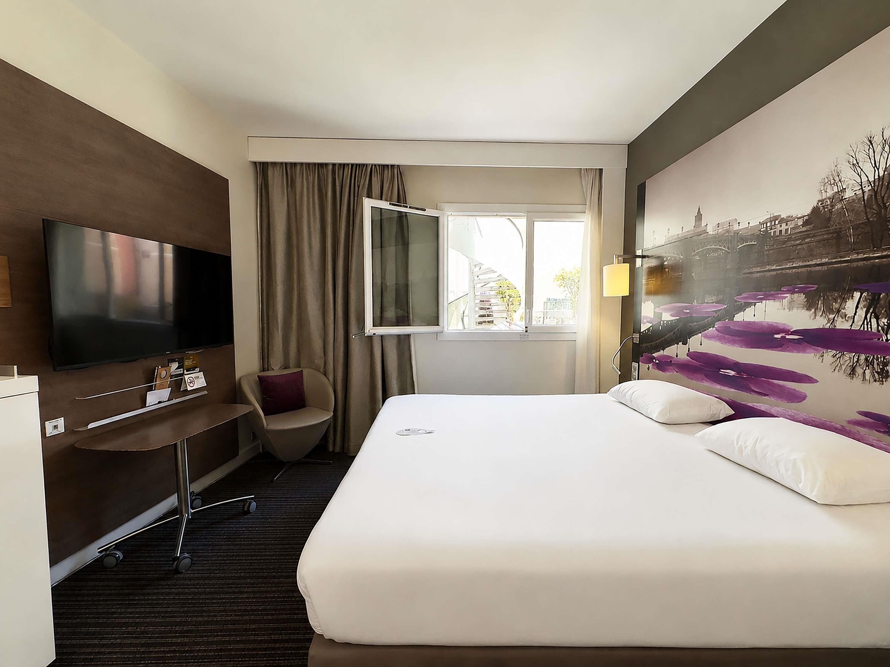A hotel room with a bed, a desk, a chair, a TV, and a window with curtains.