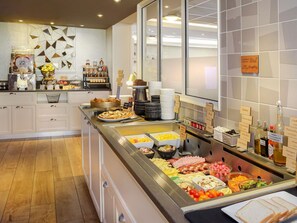 Un bufé con diversas opciones de comida y salsas, una estación de café y una pared decorativa con diseños geométricos.