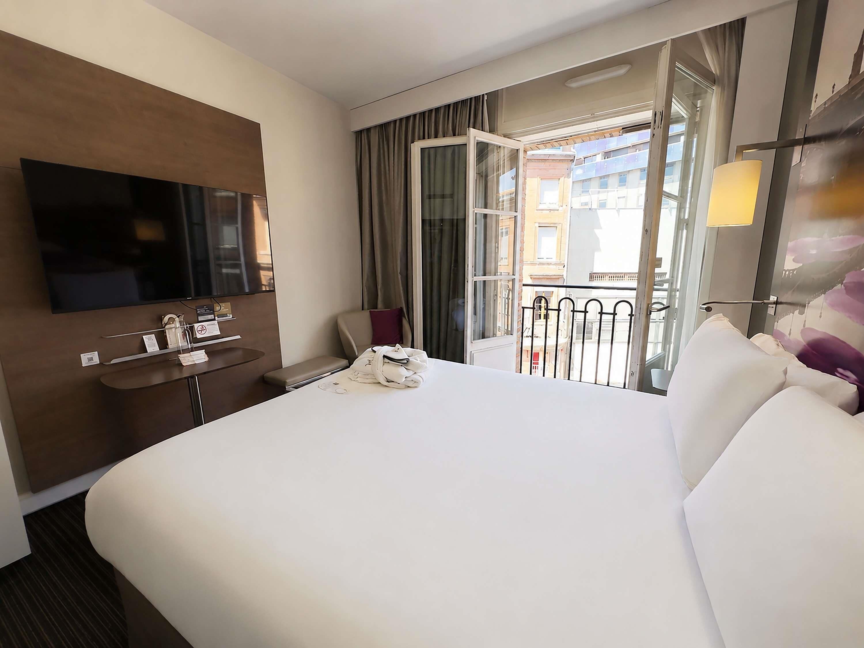 A hotel room with a bed, a TV, a desk with a phone, and a window with curtains.