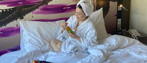 Una persona con bata blanca sentada en una cama con una bandeja de desayuno, que incluye huevos, bollería y una taza de café.