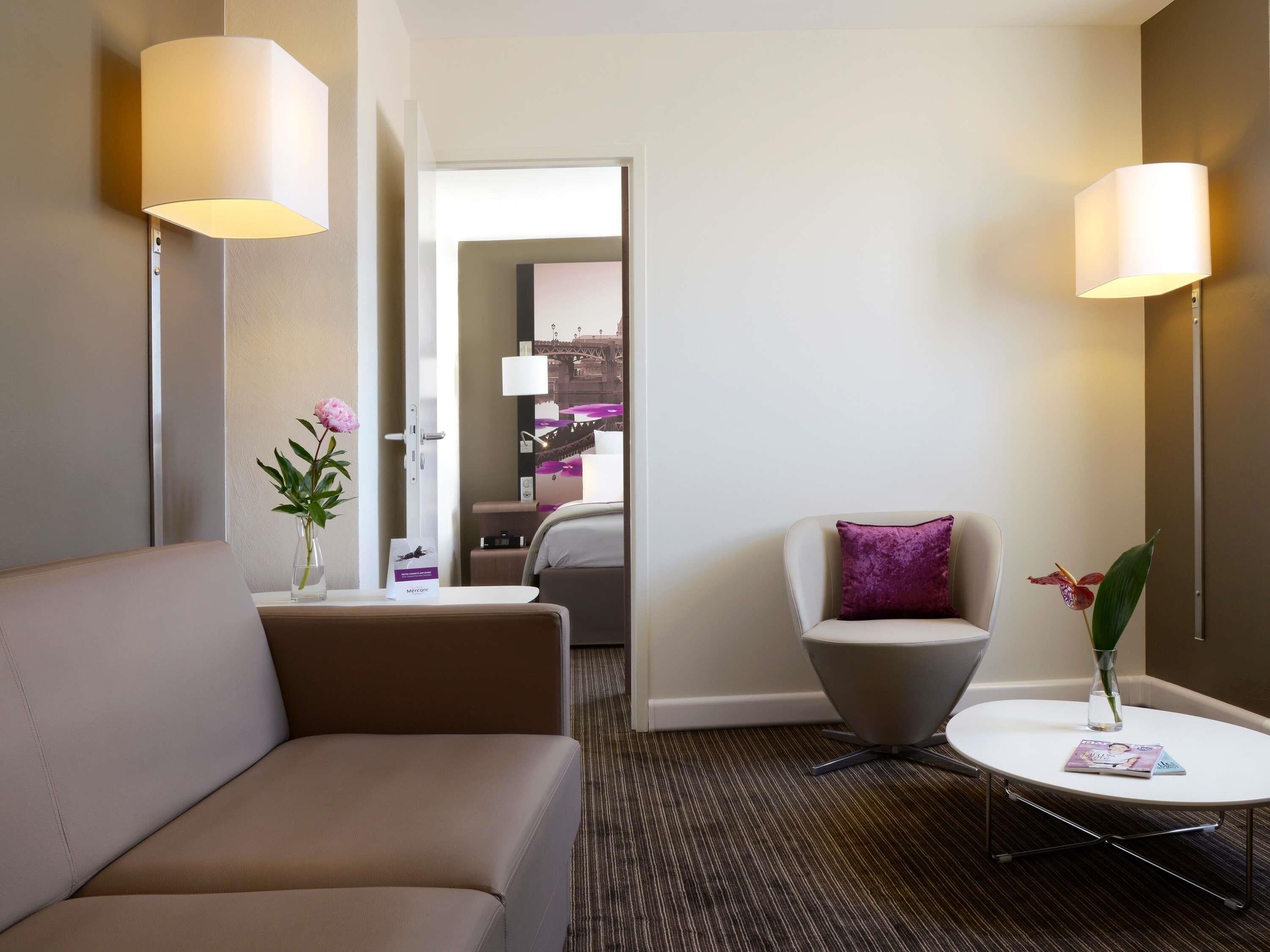 Une chambre d’hôtel avec un canapé, une petite table ronde, une chaise, un vase avec des fleurs et un lit en arrière-plan.